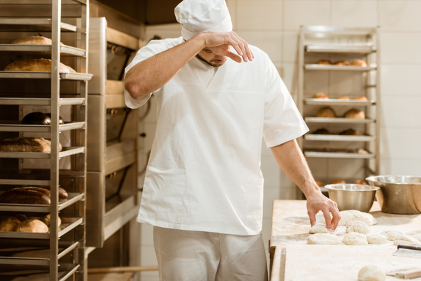 вымотанный пекарь, работающий с сырым тестом на производстве выпечки и вытирая пот
 - Фото, изображение