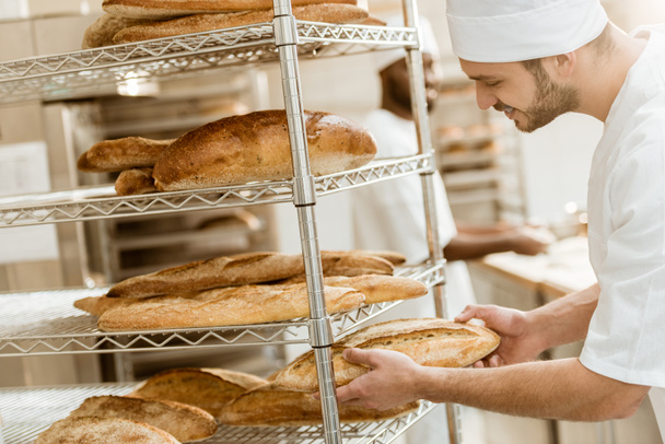 panadero joven guapo poner pan fresco en estante en la fabricación de la hornada
 - Foto, Imagen