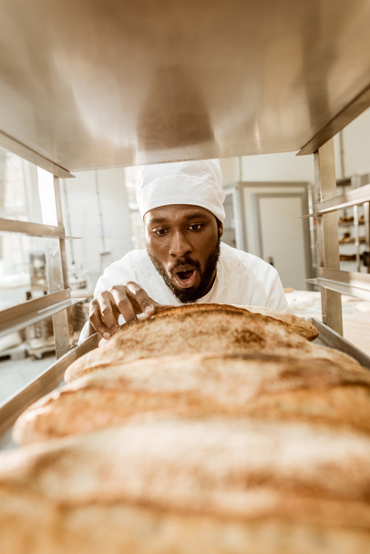 padeiro afro-americano chocado olhando para pães frescos na fabricação de panificação
 - Foto, Imagem