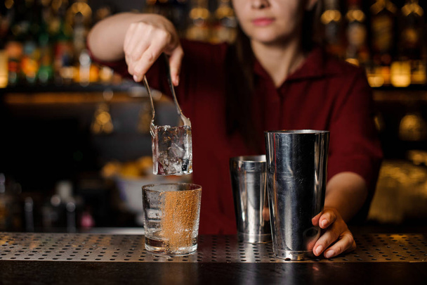 Barmen girl putting a big ice cube into an empty glass on a bar  - Fotó, kép
