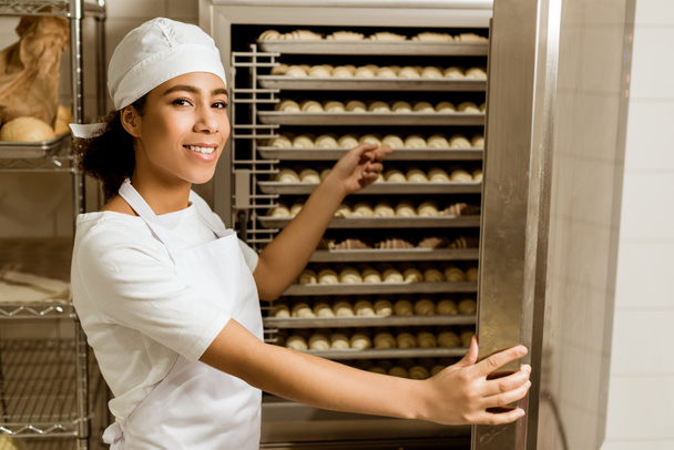 усміхнена жінка-пекар вказує на тісто всередині промислової печі на виробництві випічки
 - Фото, зображення