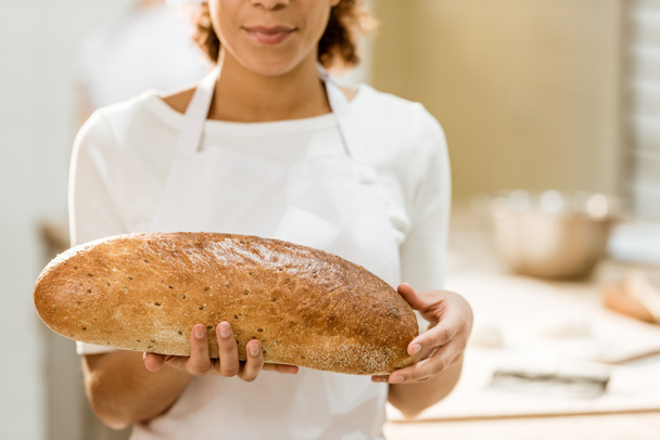 περικοπεί shot του γυναικείου baker κρατώντας το φρέσκο καρβέλι ψωμί σε ψήσιμο κατασκευή - Φωτογραφία, εικόνα