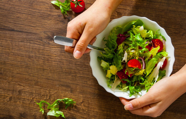 Close-up of woman hand holding various fresh mix salad leaves with tomato in bowl on wooden background on the fork  - Фото, зображення