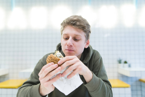 Uno studente con uno sguardo affamato guarda un hamburger tra le mani. Delizioso fast food in un ristorante leggero con un design elegante
 - Foto, immagini