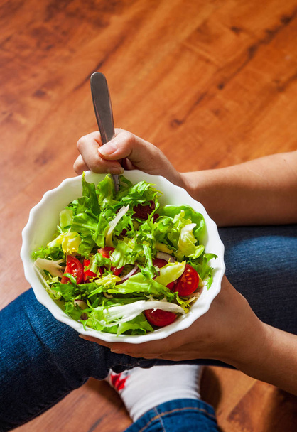 Green vegan breakfast meal in bowl with various fresh mix salad leaves and tomato. Girl in jeans holding fork with knees and hands visible, top view on wooden background - Photo, Image