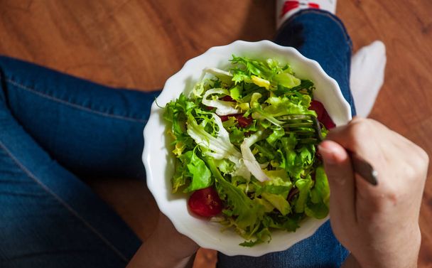 Groene veganistisch ontbijt maaltijd in kom met verschillende verse mix salade bladeren en tomaat. Meisje in spijkerbroek holding vork met knieën en handen zichtbaar, top uitzicht op houten achtergrond - Foto, afbeelding