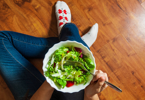 Refeição de café da manhã vegan verde em tigela com várias folhas de salada de mistura fresca e tomate. Menina em jeans segurando garfo com joelhos e mãos visíveis, vista superior sobre fundo de madeira
 - Foto, Imagem