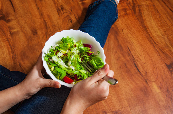 Green vegan breakfast meal in bowl with various fresh mix salad leaves and tomato. Girl in jeans holding fork with knees and hands visible, top view on wooden background - Foto, afbeelding