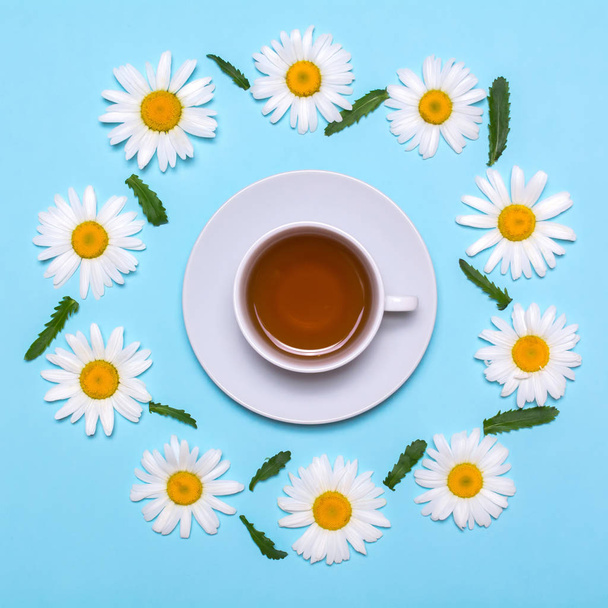 Creative layout with white tea cup with chamomile frame on a blue background - Фото, изображение