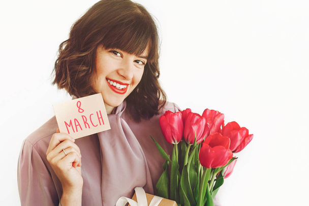 felice giorno delle donne testo sul biglietto di auguri. 8 marzo. felice ragazza elegante in possesso di carta e tulipani rosa e regalo, sorridente su sfondo bianco
.  - Foto, immagini
