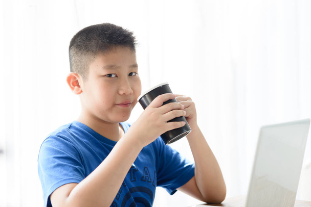 Happy Asian boy holding black cup while using tablet at home. - Фото, зображення