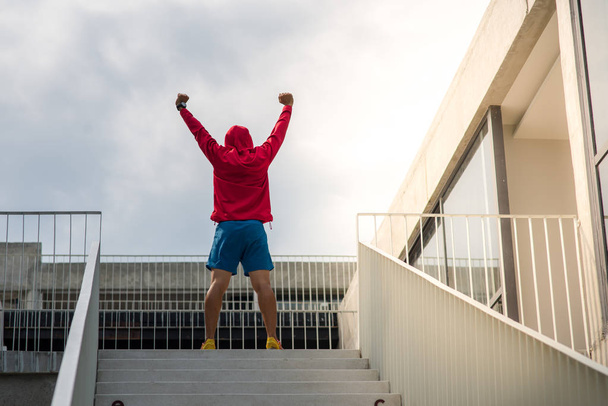 Sportman in red hood raising hands on the top of the steps outdo - Fotoğraf, Görsel
