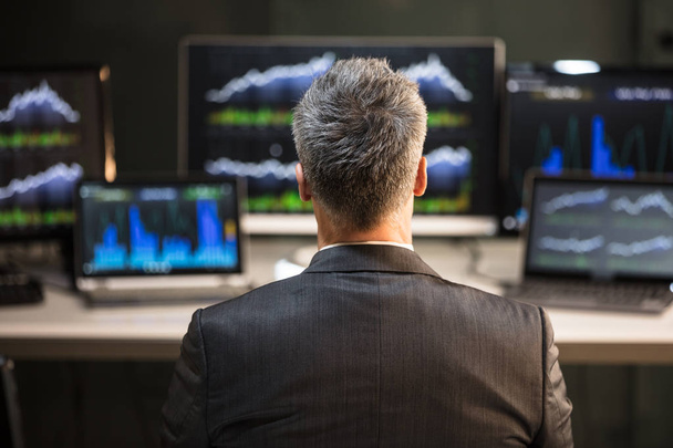Male Stock Market Broker Looking At Computer And Laptop Showing Graph On Screen - 写真・画像