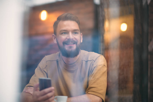 Nuori mies istuu kahvilassa ja käyttää älypuhelinta
 - Valokuva, kuva