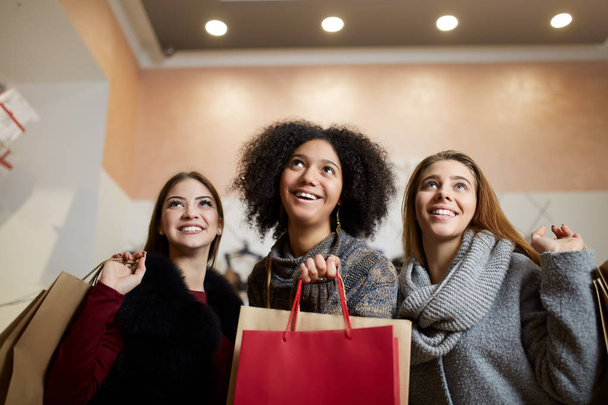 Frauen verschiedener ethnischer Zugehörigkeit mit Einkaufstüten, die in Einkaufszentren zum Verkauf stehen. Porträt von drei lächelnden multirassischen Mädchen, die auf den Copyspace-Bereich über ihren Köpfen nach Designern schauen. - Foto, Bild
