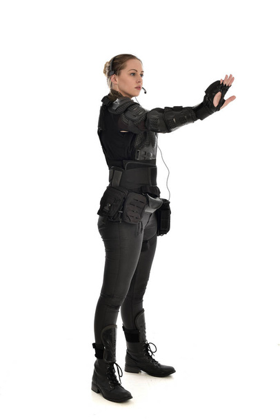 портрет женщины-солдата в черных тактических доспехах, стоящей в профиль, изолированной на белом фоне студии
. - Фото, изображение