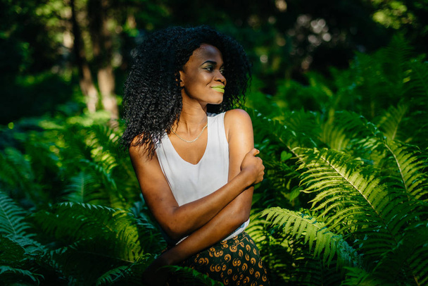 平和的に笑みを浮かべて、シダの中で立ちながらよそ見緑のメイクアップと非常に美しいアフリカの女の子の柔らかい屋外のポートレート. - 写真・画像