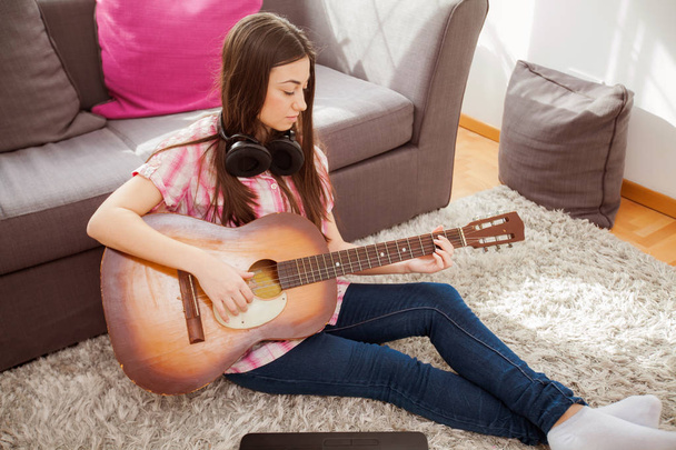 Mujer joven tocando la guitarra acústica, sentada en el suelo con ropa casual en casa
 - Foto, Imagen