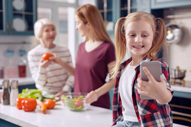 Smiling girl taking selfie while mother and grandmother cooking - Zdjęcie, obraz