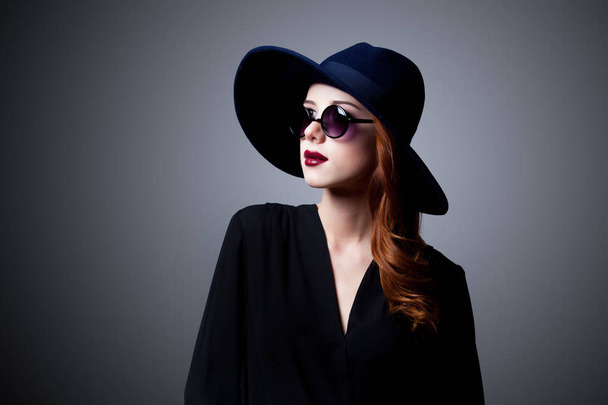 Young redhead girl in dark style in sunglasses and hat on grey background  - Photo, Image