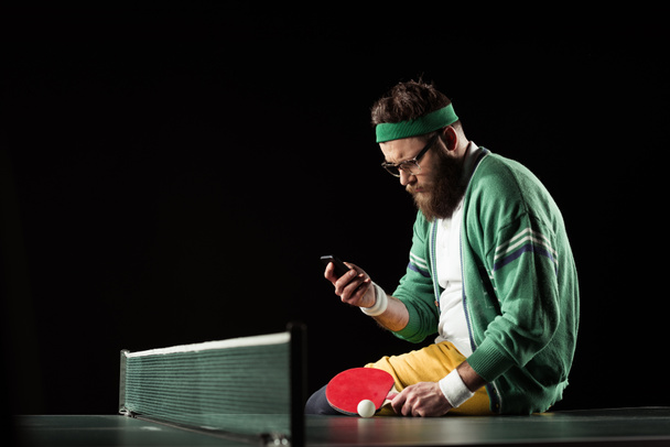 tennis player using smartphone while sitting on tennis table isolated on black - Фото, зображення