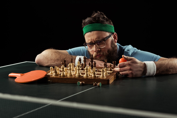 homem barbudo pensivo olhando para tabuleiro de xadrez na mesa de tênis isolado em preto
 - Foto, Imagem