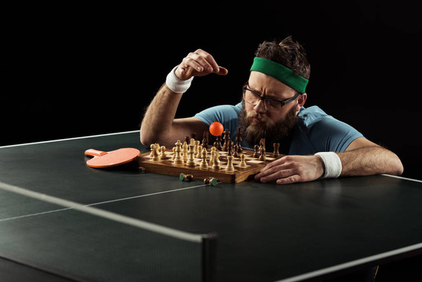 konzentrierter bärtiger Tennisspieler wirft Ball auf Schachbrett auf Tischtennisplatte isoliert auf schwarz - Foto, Bild