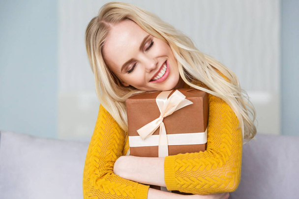 belle fille blonde heureuse avec les yeux fermés étreignant boîte cadeau à la maison
 - Photo, image