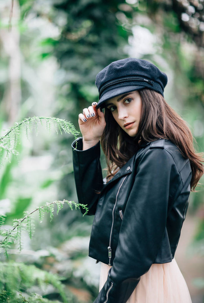 Fashion portrait of young stylish woman in leather jacket and cap on nature background. Young model poses near cacti, holds visor with fingers - 写真・画像