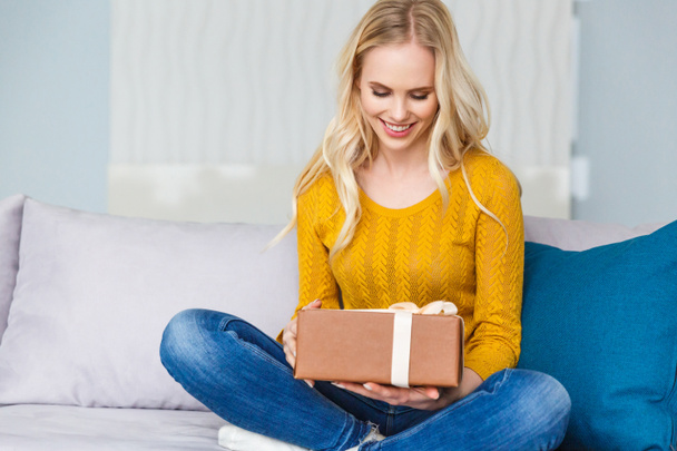 красива усміхнена молода жінка тримає подарункову коробку, сидячи на дивані вдома
 - Фото, зображення