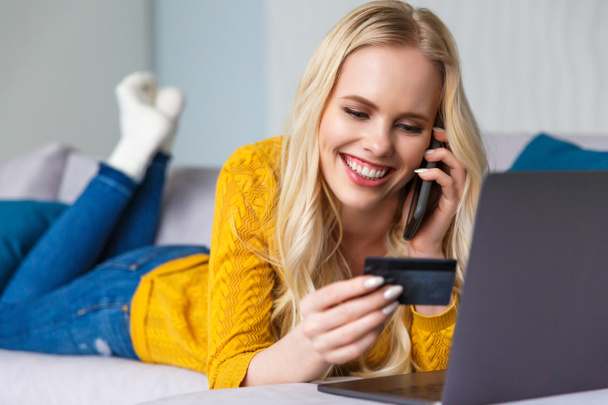 beautiful smiling young woman holding credit card and talking by smartphone at home - Photo, Image