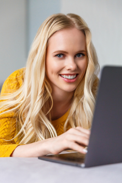 όμορφη νεαρή ξανθιά γυναίκα χρησιμοποιώντας φορητό υπολογιστή και να χαμογελά στη φωτογραφική μηχανή - Φωτογραφία, εικόνα