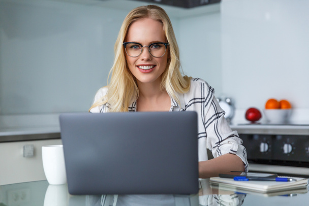 beautiful young woman in eyeglasses smiling at camera while working with laptop at home - Photo, Image
