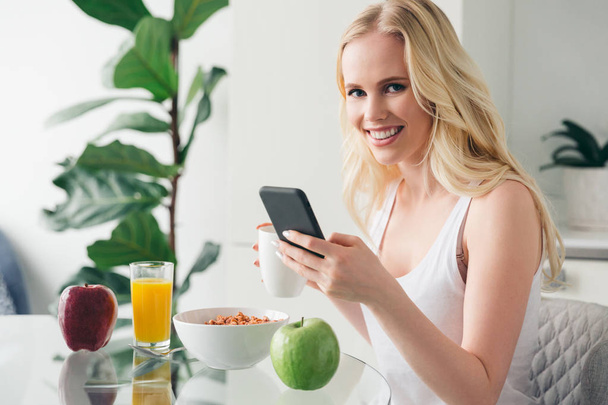 gyönyörű fiatal nő használ smartphone és mosolyogva kamera otthon reggelire - Fotó, kép