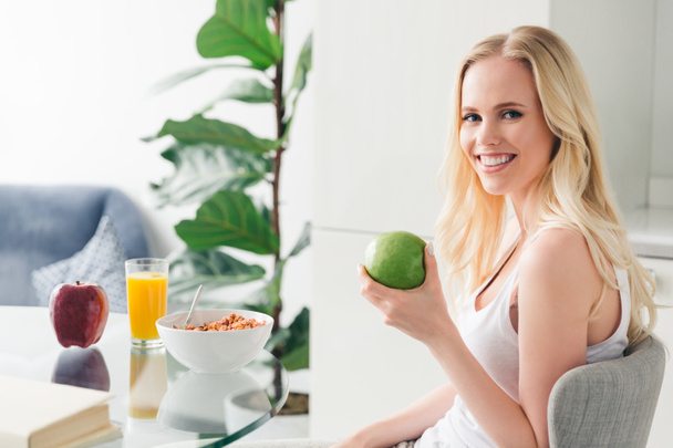 beautiful blonde girl holding apple and smiling at camera while having breakfast at home - Photo, Image