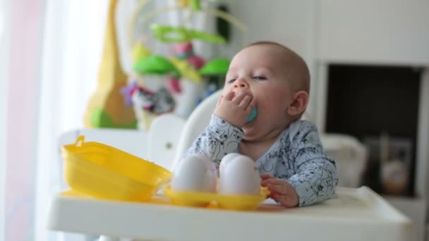 Cute little toddler boy, playing with plastic eggs, sitting in a white chair in a sunny living room, white around him, baby boy smiling happily - Felvétel, videó