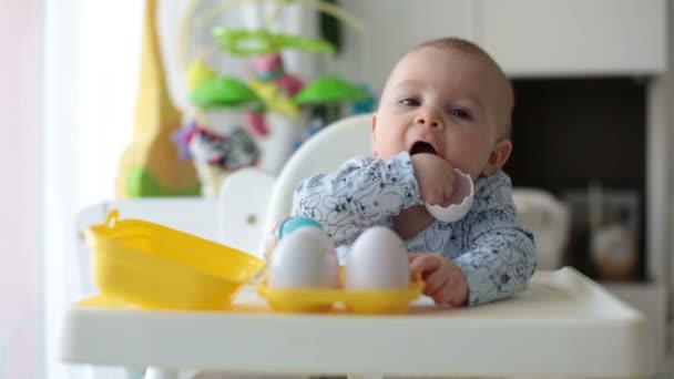Cute little toddler boy, playing with plastic eggs, sitting in a white chair in a sunny living room, white around him, baby boy smiling happily - Séquence, vidéo