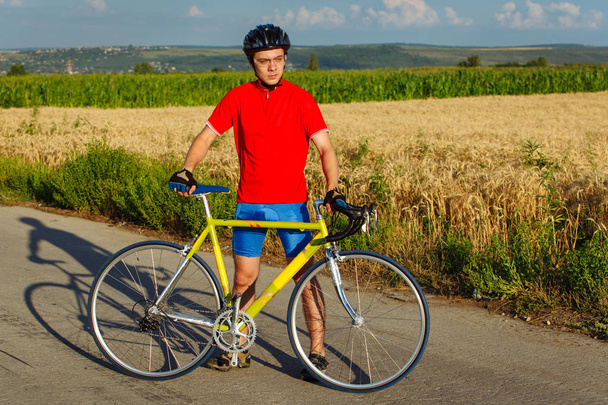 Urheilija seisoo polkupyörän vieressä. Yllään urheiluvälineet, kypärä ja lasit. Luonnollinen tausta
. - Valokuva, kuva