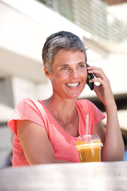 Retrato de una mujer mayor feliz sentada afuera con bebida y hablando por teléfono móvil
 - Foto, Imagen