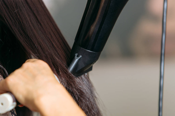 Gros plan de coiffeurs mains séchant de longs cheveux noirs avec sèche-cheveux et brosse ronde
. - Photo, image