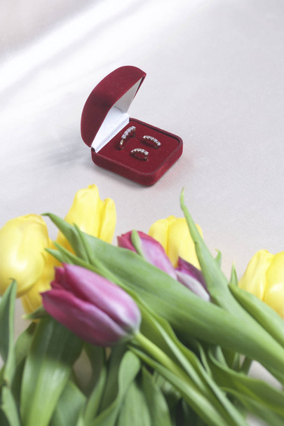 Gifts for loved ones. A bouquet of yellow and pink tulips is scattered on a light surface. Nearby is an open velvet box of red color with gold jewelry. - Foto, Imagem