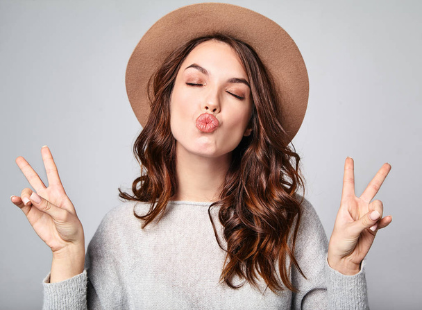 Portrait of young stylish laughing girl model in gray casual summer clothes in brown hat with natural makeup isolated on gray background. Looking at camera and showing peace sign and giving air kiss - Foto, immagini