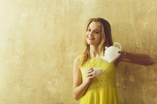 happy blonde girl in yellow dress holds cup and teapot - Fotoğraf, Görsel