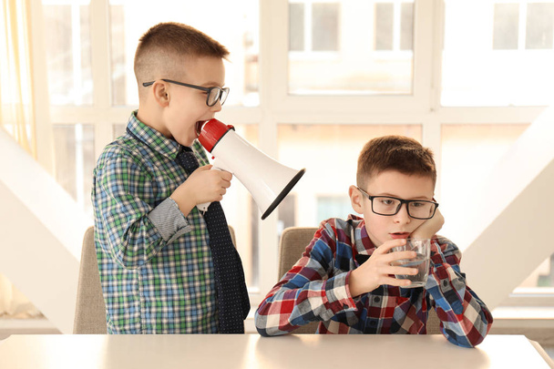 Funny little boy with megaphone shouting at his bored friend sitting at table - Foto, Imagen