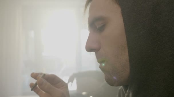 Close up of a young male reggae musician in hood sitting on the couch with his friend smoking weed on the couch in his rented apartment - Footage, Video