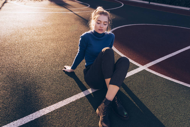 stylish young blond model girl in blue sweater posing on sports field in the morning sun, street style and fashion - Фото, изображение