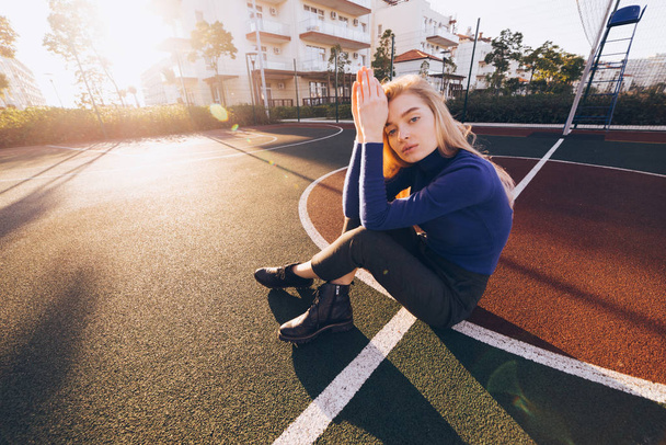 stylish young blond model girl in blue sweater posing on sports field in the morning sun - Foto, Imagem