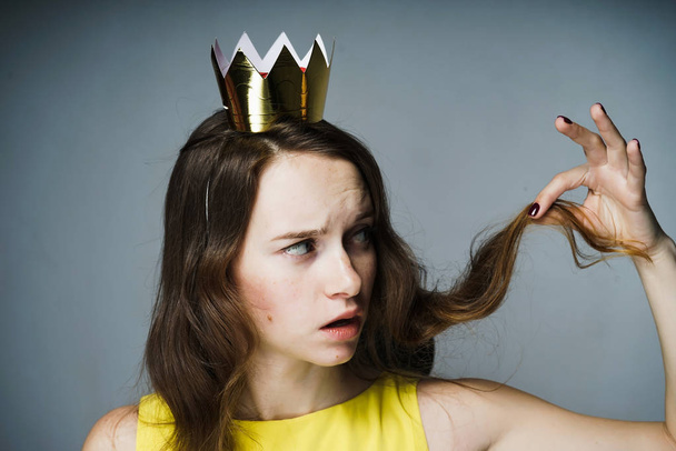 dissatisfied young girl holding a curl of hair, on her head a golden crown - Fotoğraf, Görsel