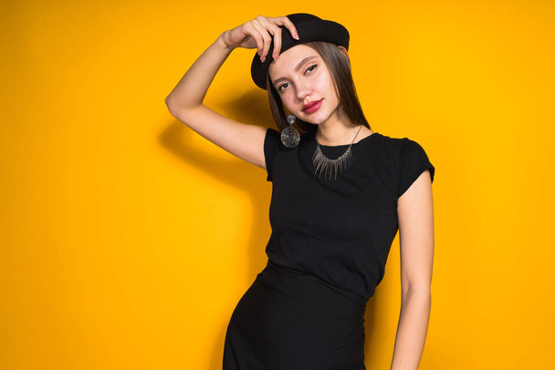 confident beautiful girl in black dress and fashionable hat posing on orange background, smiling - Photo, Image