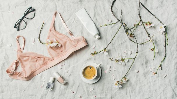 Woman's morning rituals concept. Feminine tender pink lingerie, glasses, cosmetic items, cup of coffee and Spring almond blossom flowers over light grey linen bed cover, top view. - Zdjęcie, obraz
